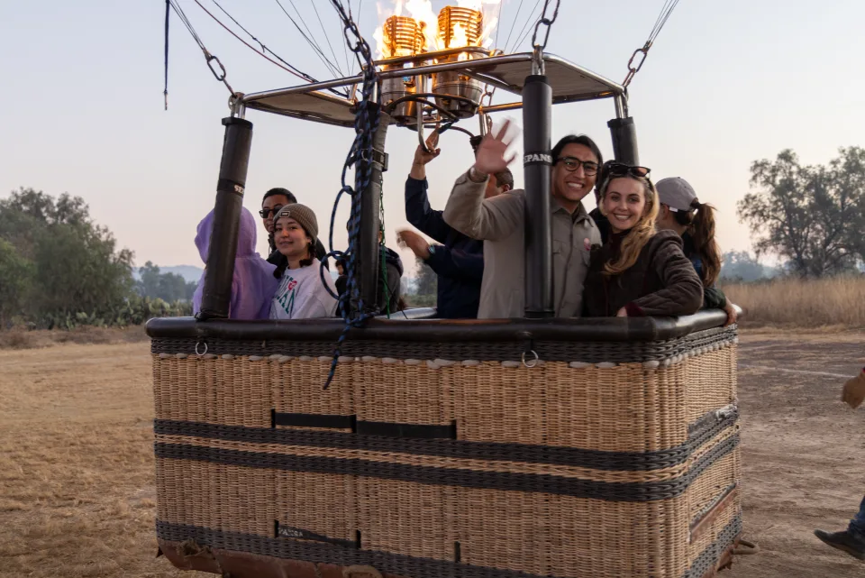 Real passengers waving from a hot air balloon basket before a sunrise flight over Teotihuacan